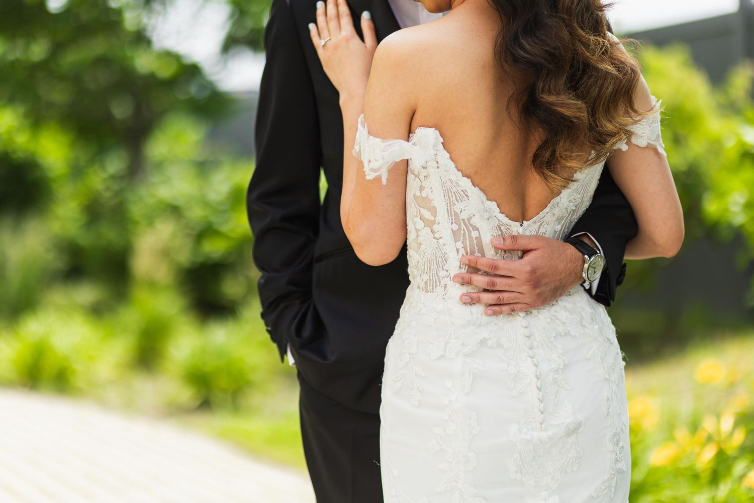 A tender moment between newlyweds showing the groom's hand on the bride's lace dress against a green outdoor backdrop.