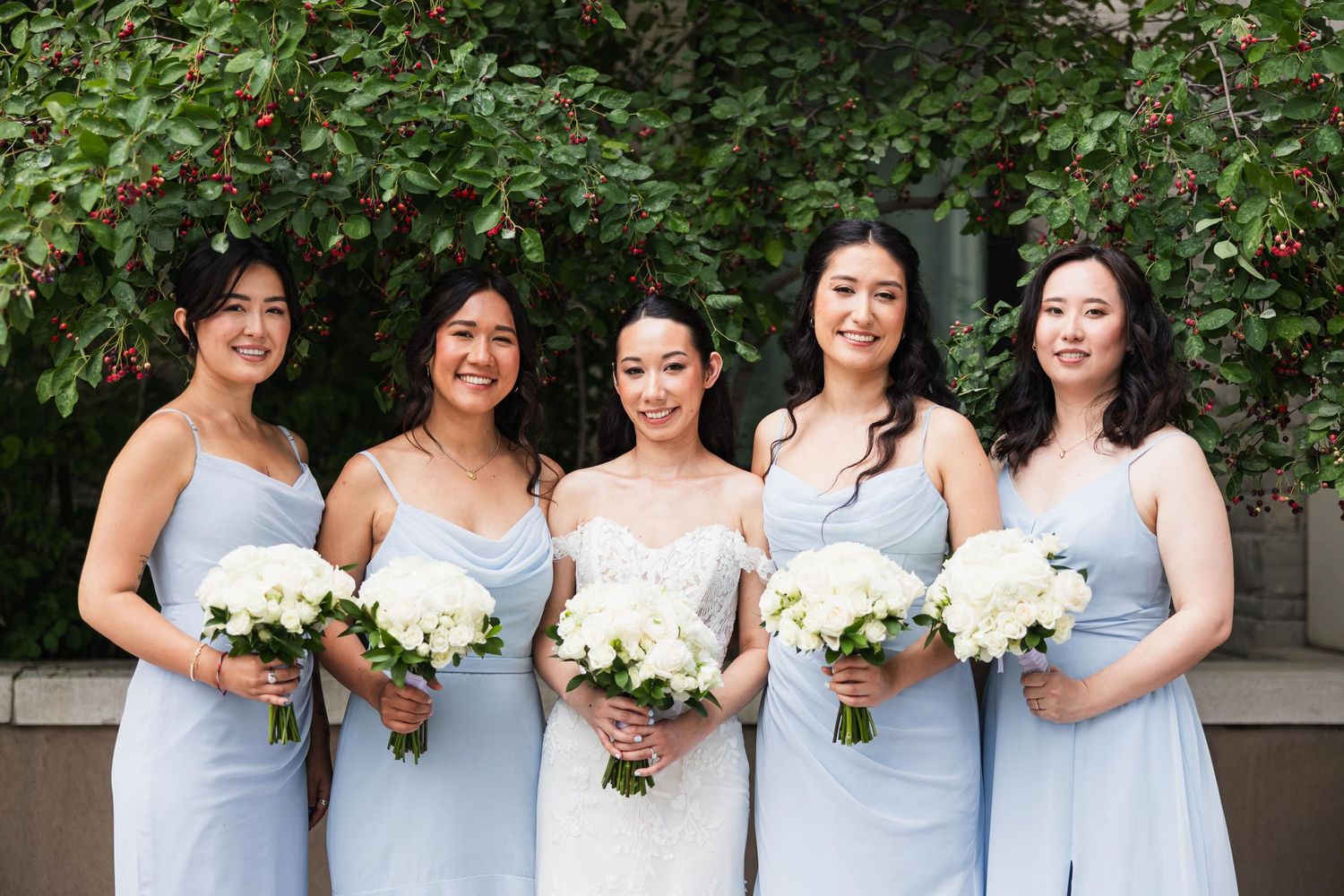 Bridesmaids in light blue dresses hold white bouquets while posing outdoors.