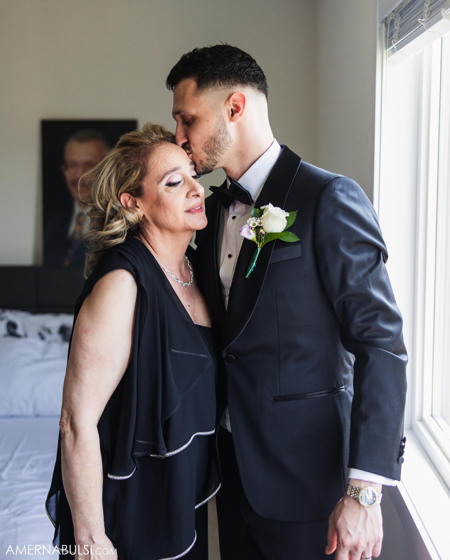 Couple shares tender embrace near window while wearing formal black attire and floral boutonniere.