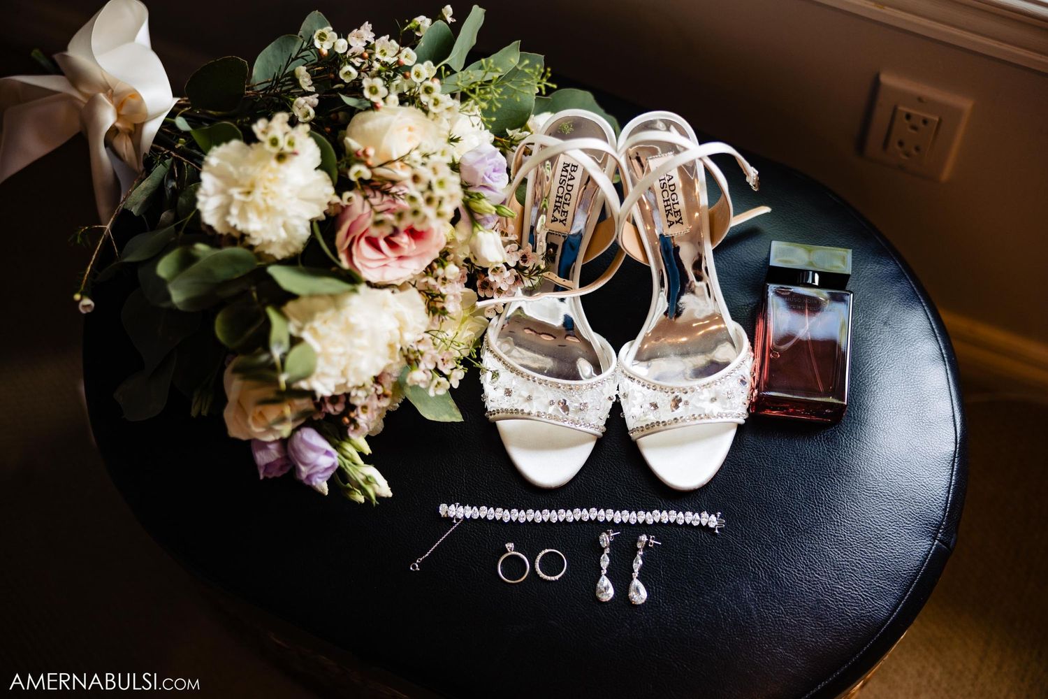 Wedding accessories including white heels, bouquet, perfume and jewelry arranged on dark round table.