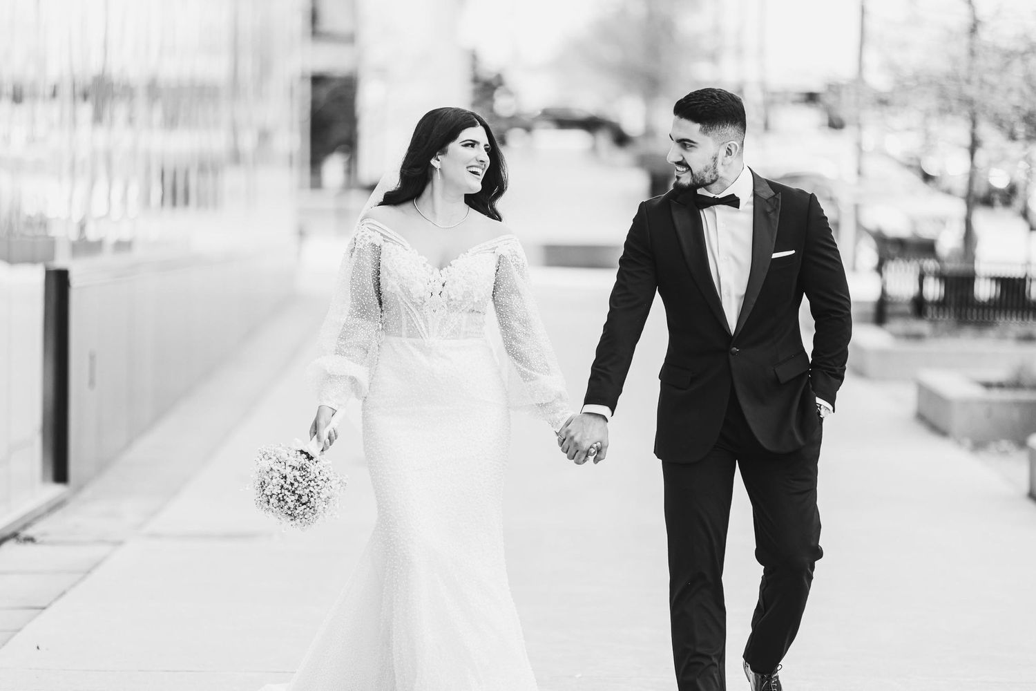 Black and white photo of newlyweds walking together, bride holding bouquet in lace-sleeved gown.