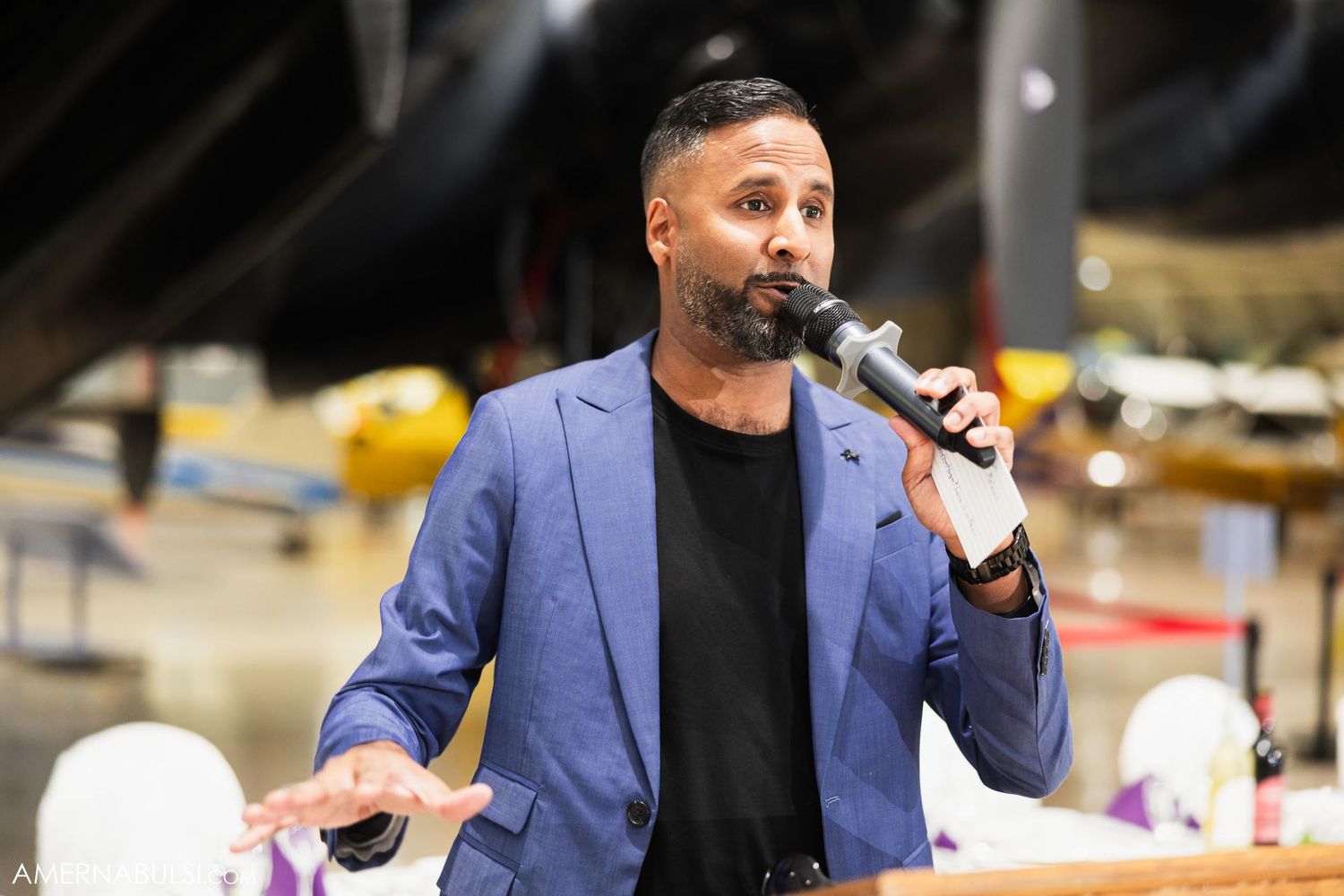 A speaker in a blue blazer and black shirt presents at an event with a microphone.