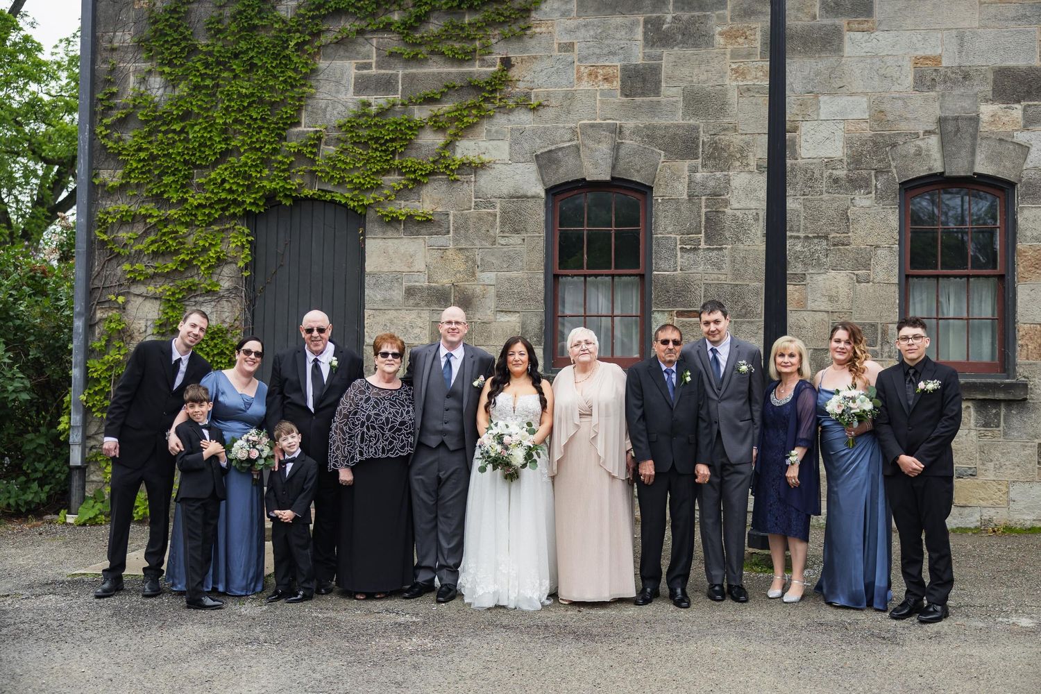 Large family wedding portrait with guests in formal attire gathered outside historic stone building.