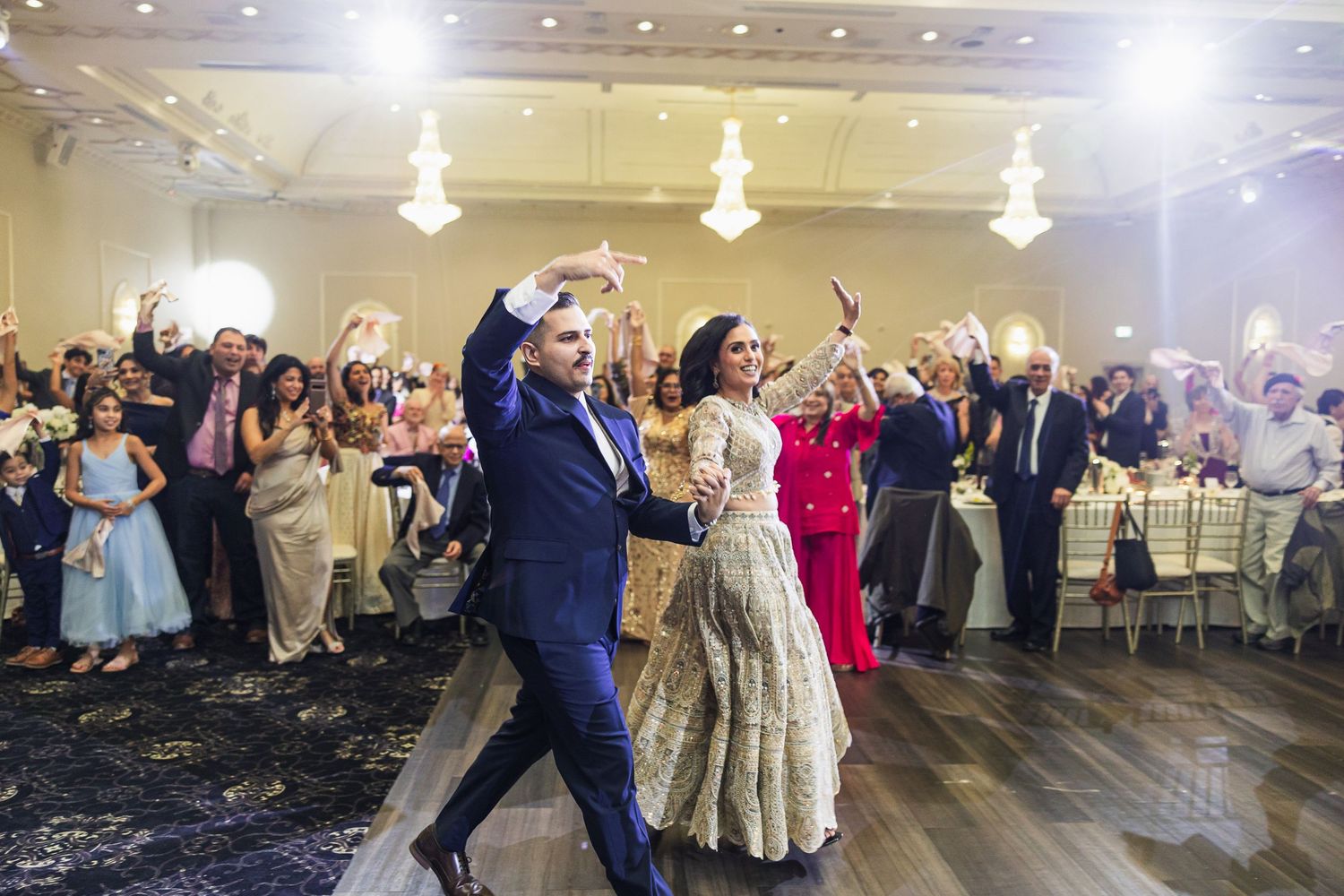 Large group of wedding guests dancing together in an elegant ballroom with chandeliers.