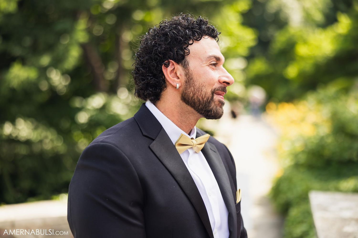 Profile view of groom in black suit and gold bowtie during outdoor wedding ceremony.