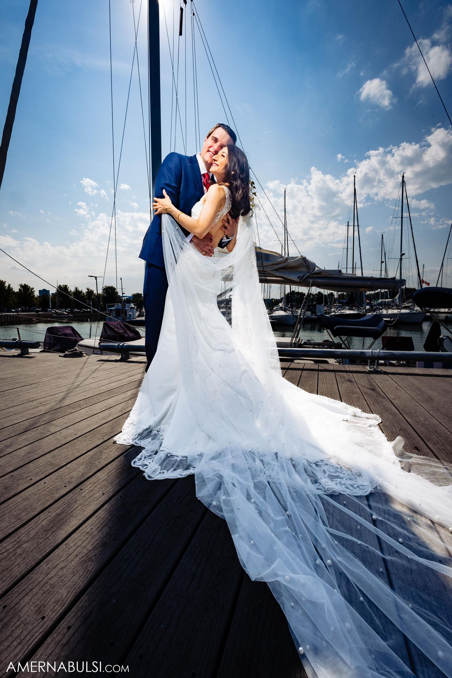 A stunning wedding photo of a couple embracing on a marina dock, with the bride's lace gown and veil trailing dramatically.