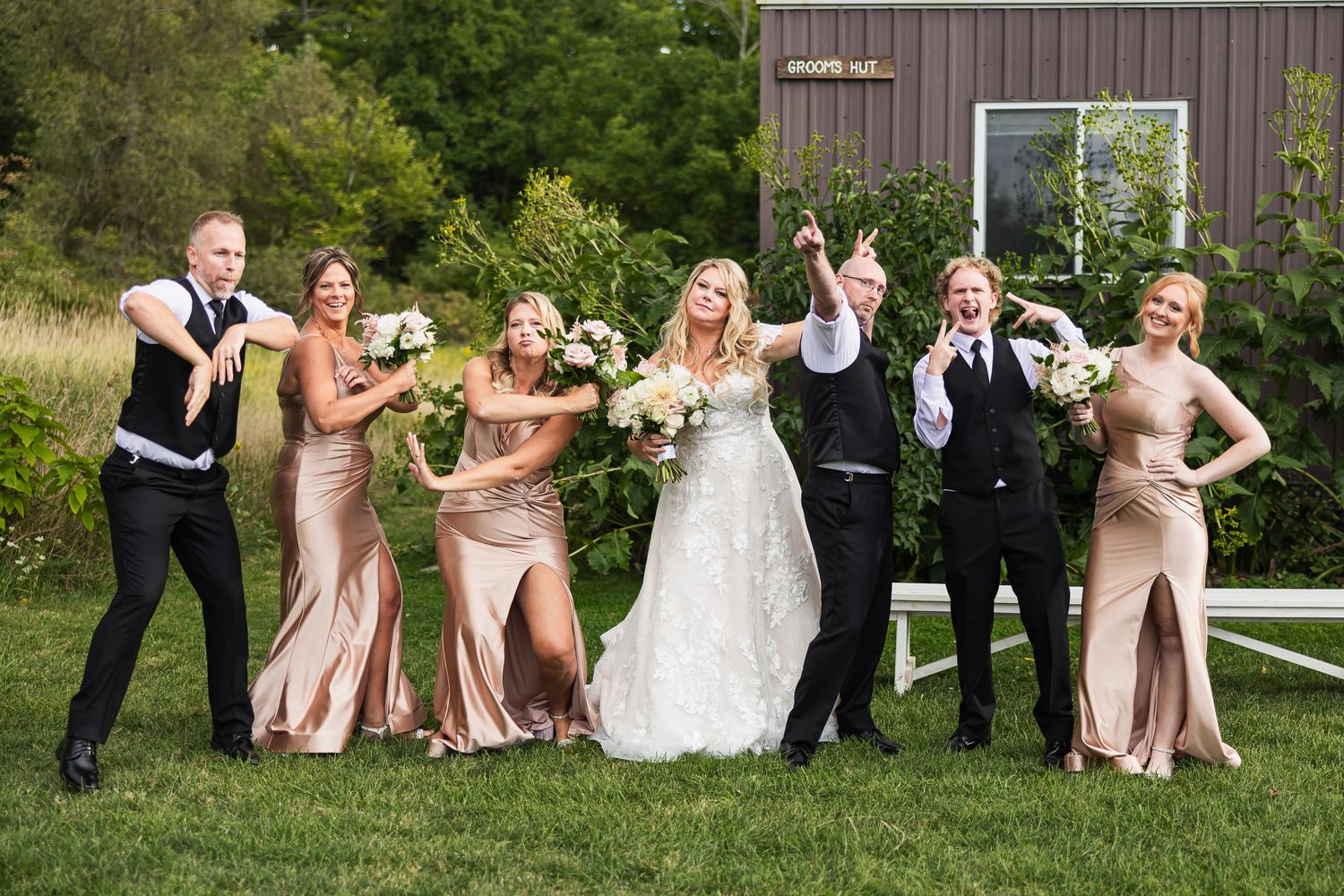 Wedding party members in pink dresses and black suits playfully pose in front of wooden barn.