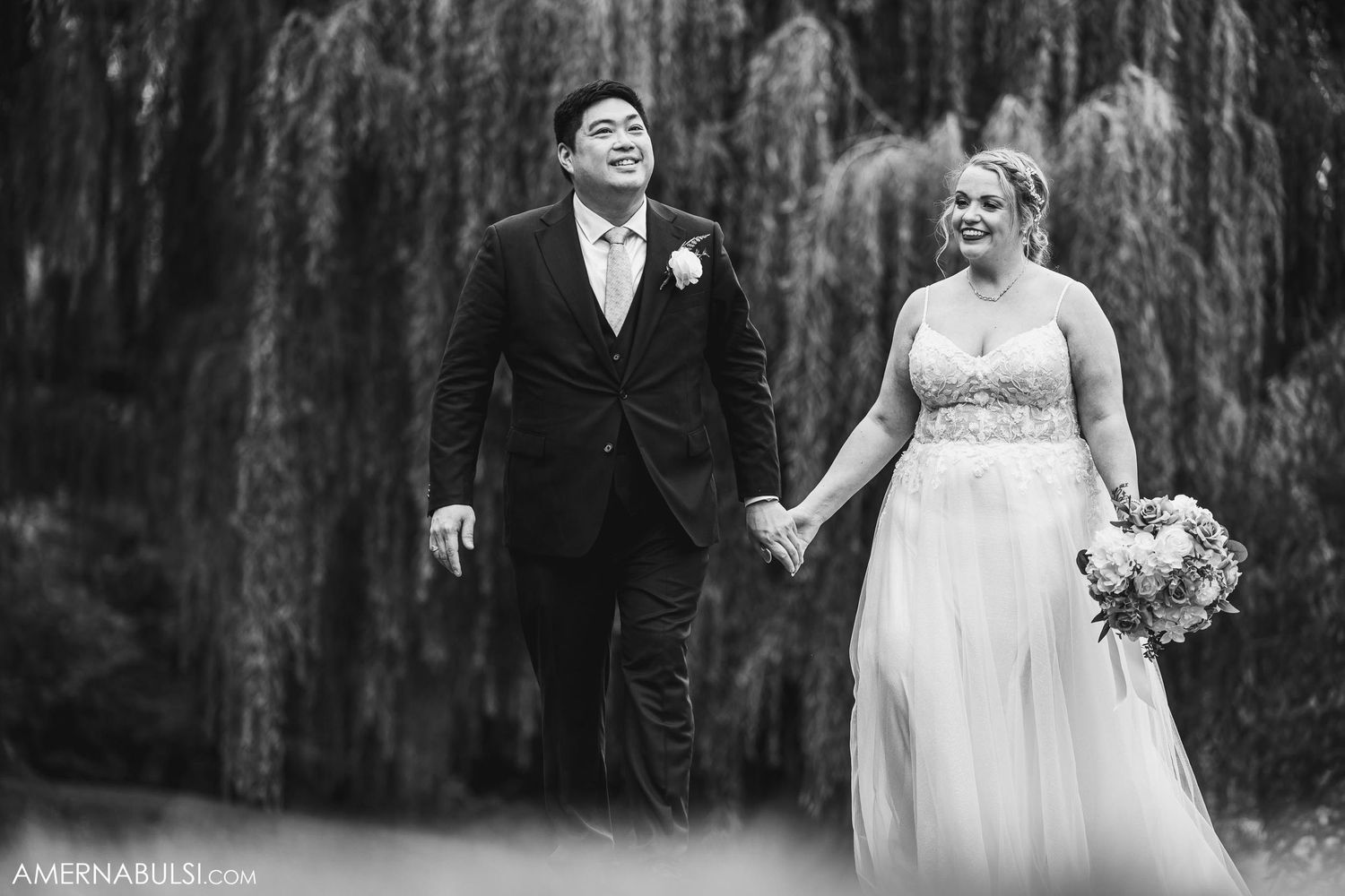 A romantic black and white wedding photo featuring a couple walking under weeping willow trees.