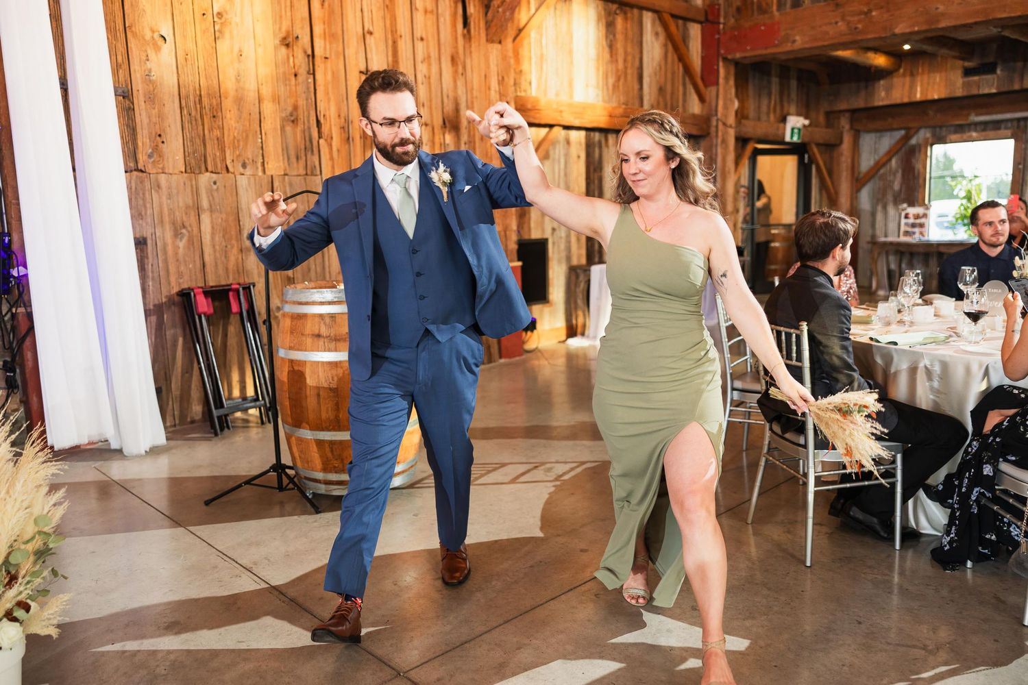Wedding party members dance energetically in a rustic barn reception venue.