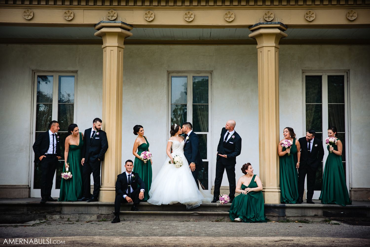 Wedding party in formal attire poses along a building exterior with emerald green dresses and classic columns.