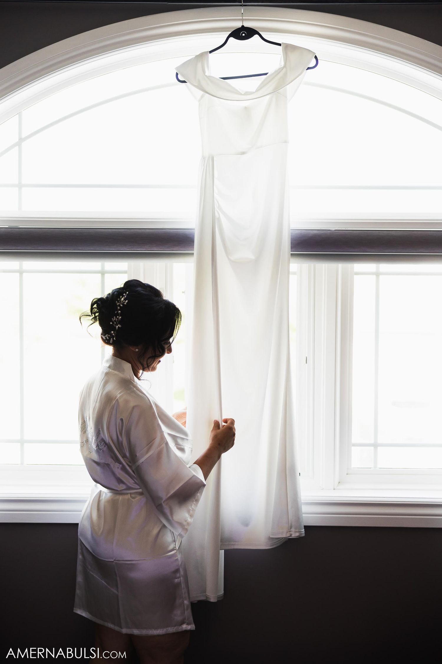 Simple white wedding gown hanging in front of an arched window.
