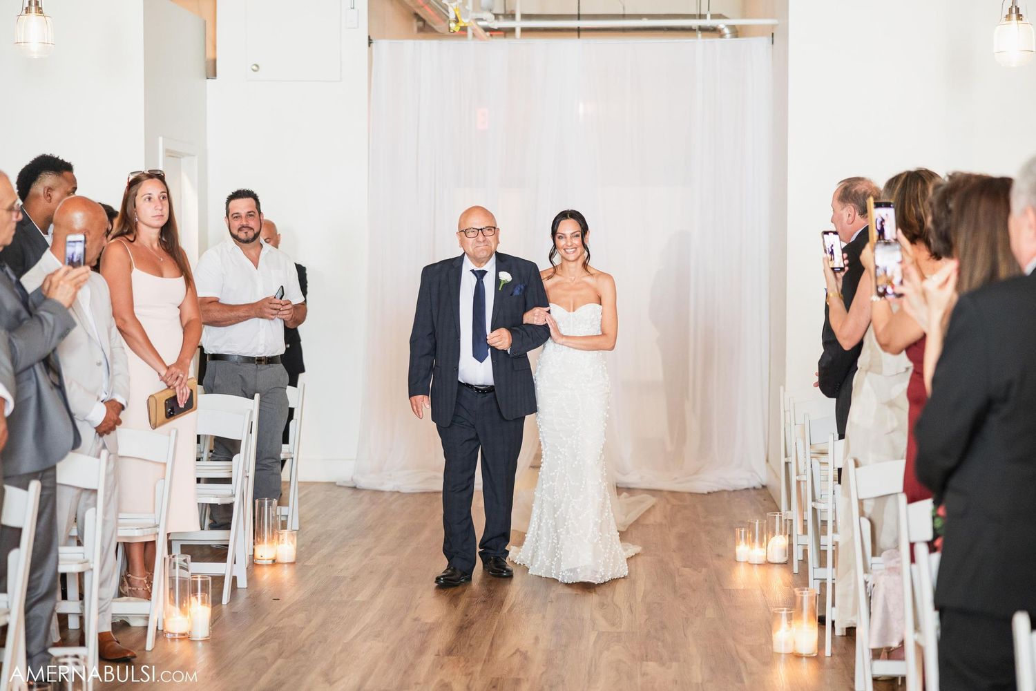 Father walks bride down candle-lit aisle in modern industrial wedding venue.