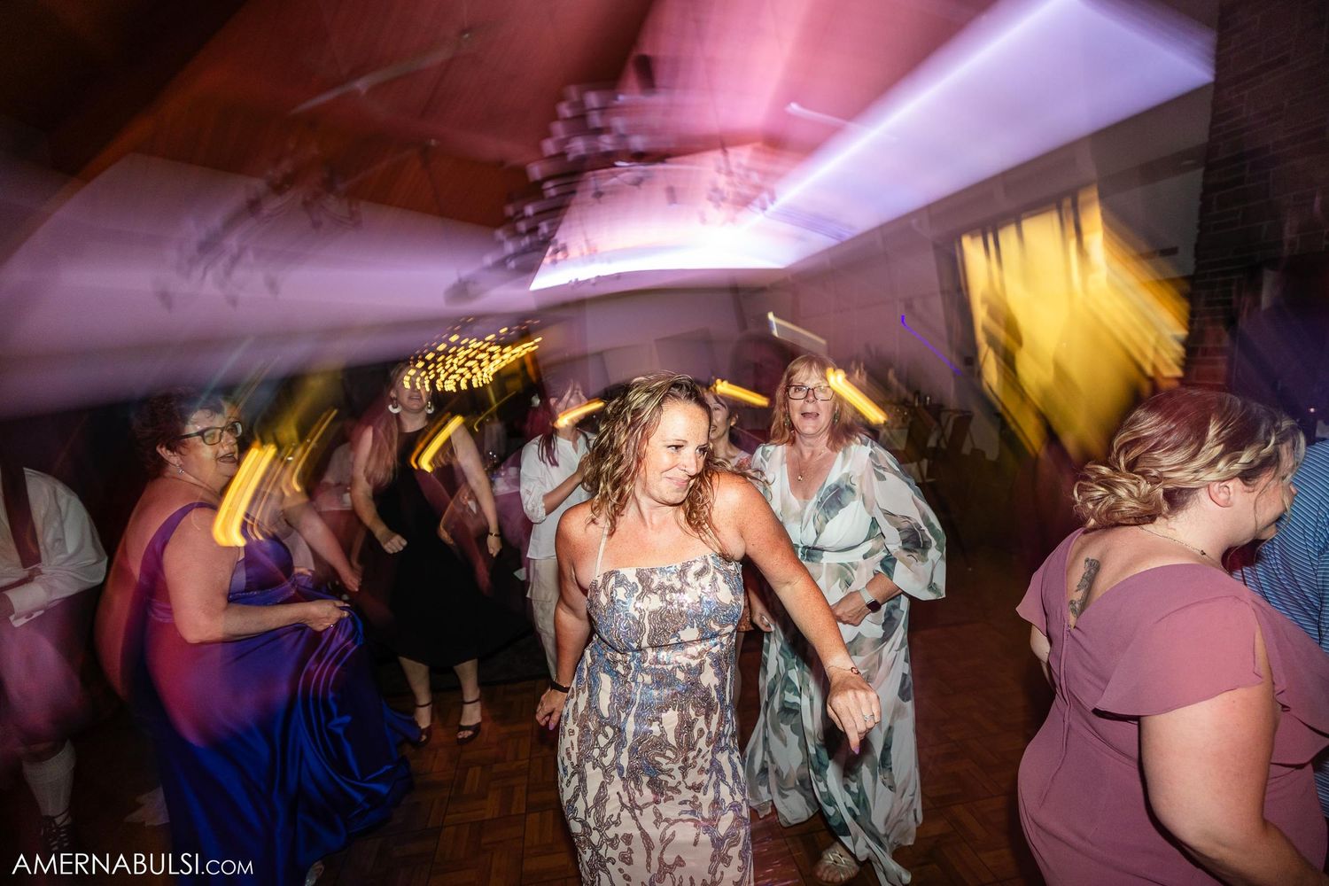 Wedding guests dancing energetically on a dance floor under pink lighting at a reception celebration.