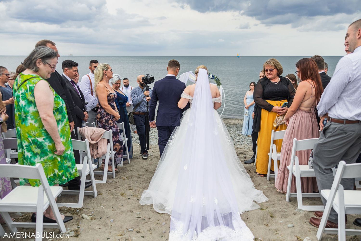 Beach wedding ceremony with guests seated in white chairs along the shoreline under cloudy skies.