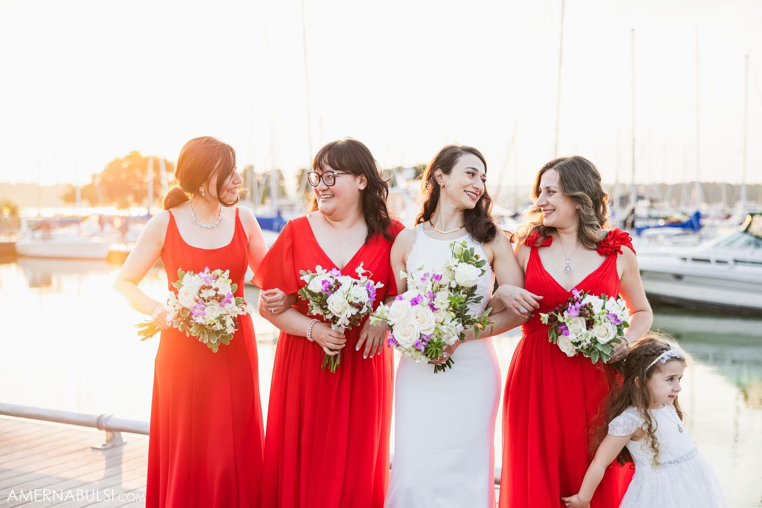 Bridesmaids in bright red dresses hold bouquets against sunset marina backdrop.