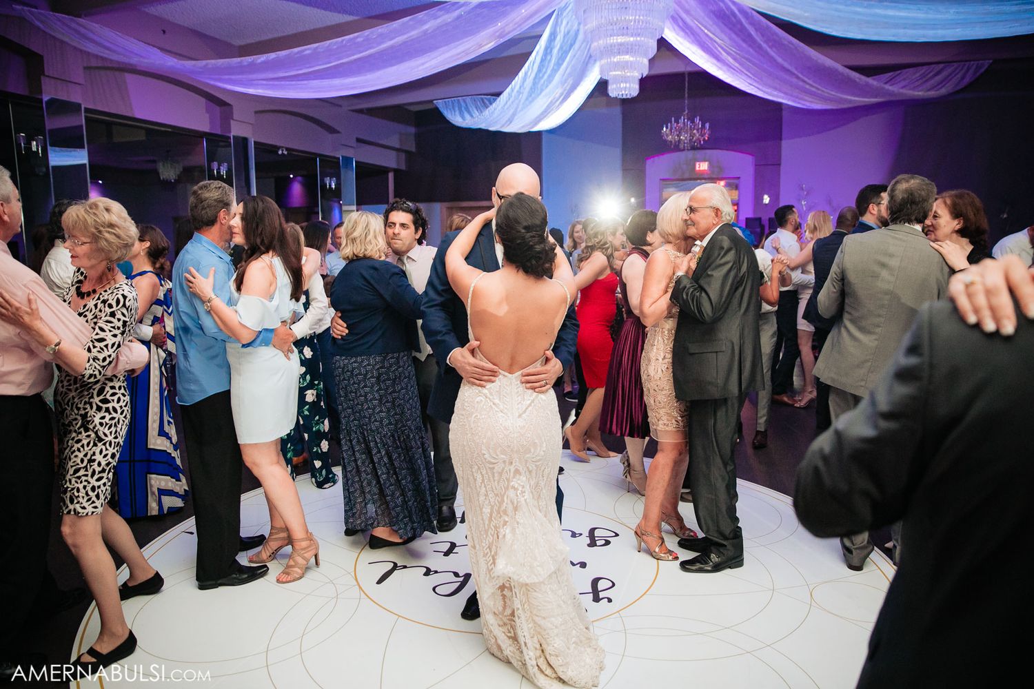 Panoramic view of a wedding reception dance floor with purple uplighting and white ceiling draping.