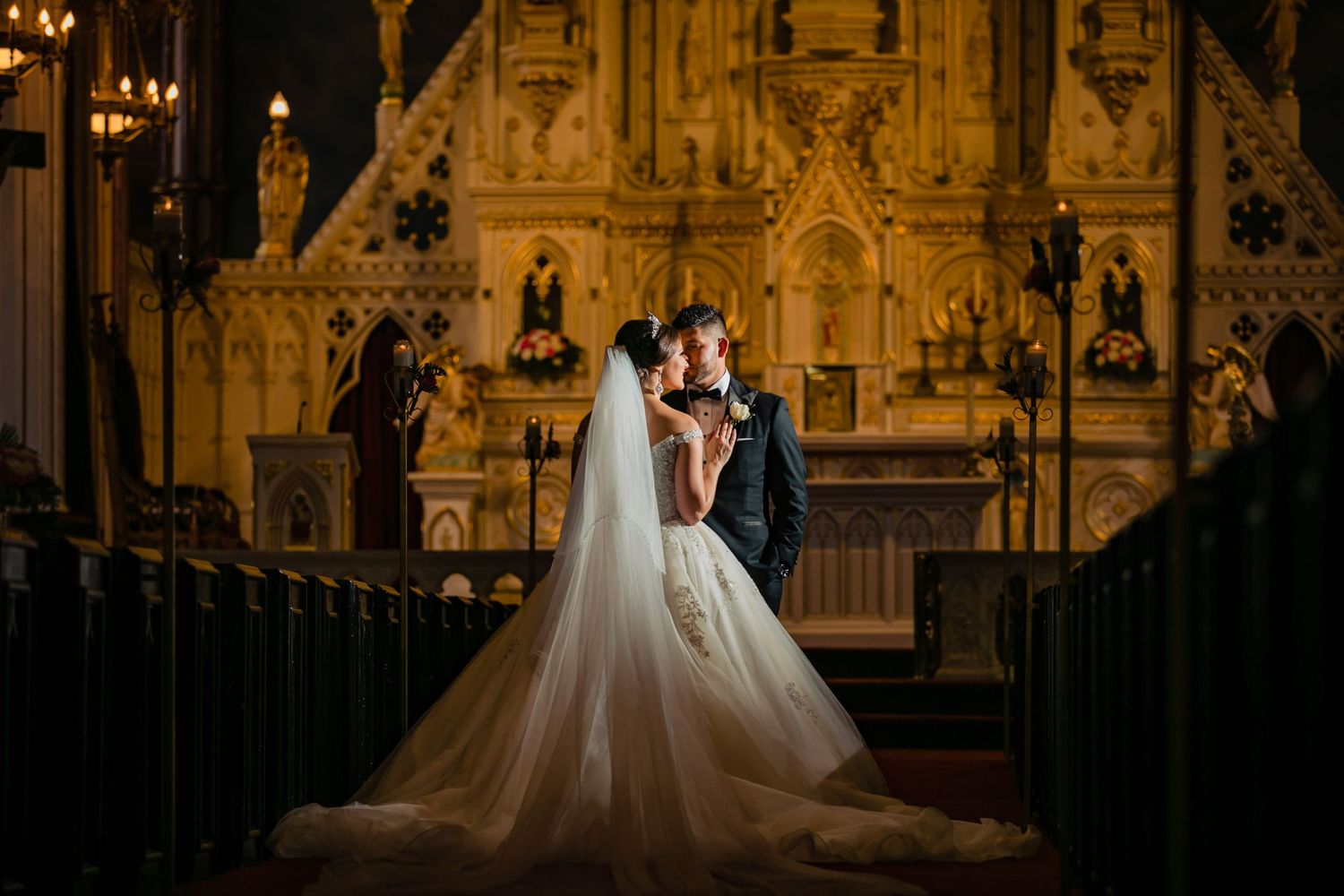 A couple in wedding attire stands silhouetted in a grand cathedral with ornate Gothic architecture and glowing lights.