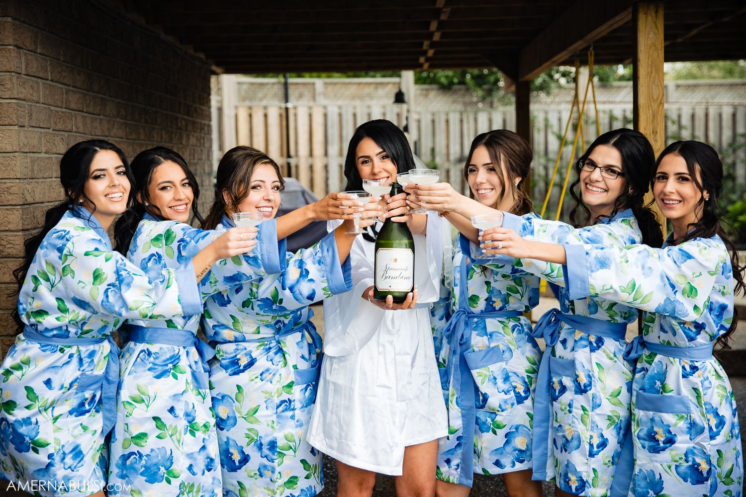 Group in matching blue floral robes and one in white dress toast with champagne glasses during celebration event.