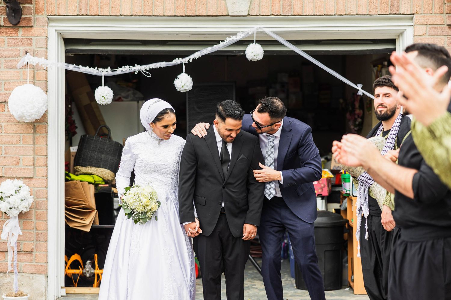 A wedding party exits a brick building decorated with white lanterns as guests applaud.