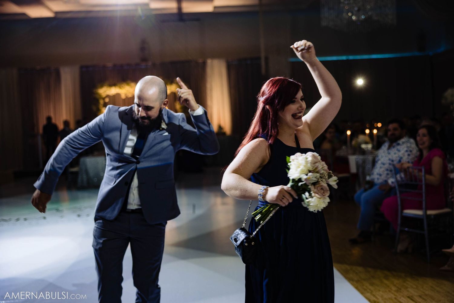Wedding party members dance energetically while holding bouquets at an indoor reception.