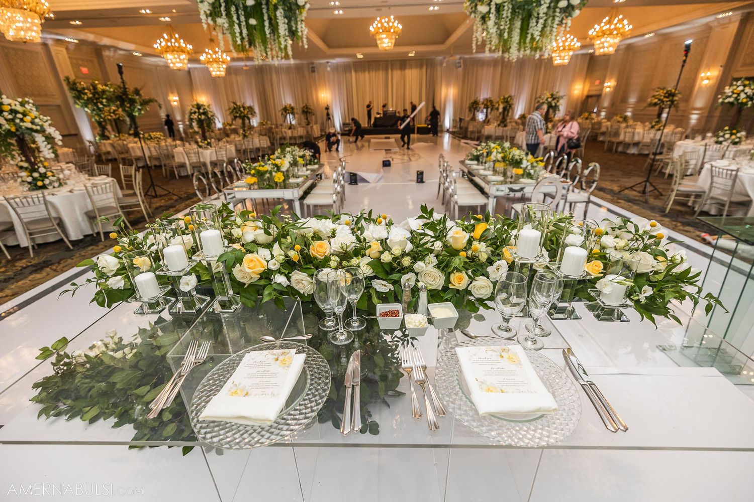 Elegant wedding reception table with white and yellow floral centerpiece arrangements and reflective mirrored surface.