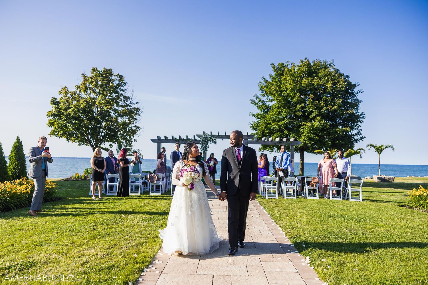 An outdoor wedding ceremony unfolds on a grassy hill overlooking a scenic waterfront vista.