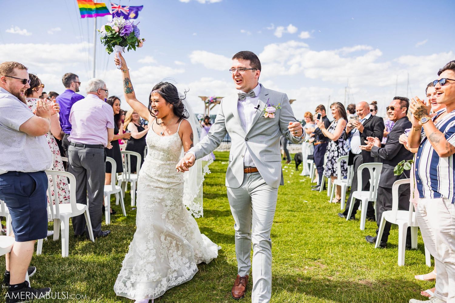 Newlyweds walk down aisle between white chairs as guests celebrate under blue sky with rainbow flag.