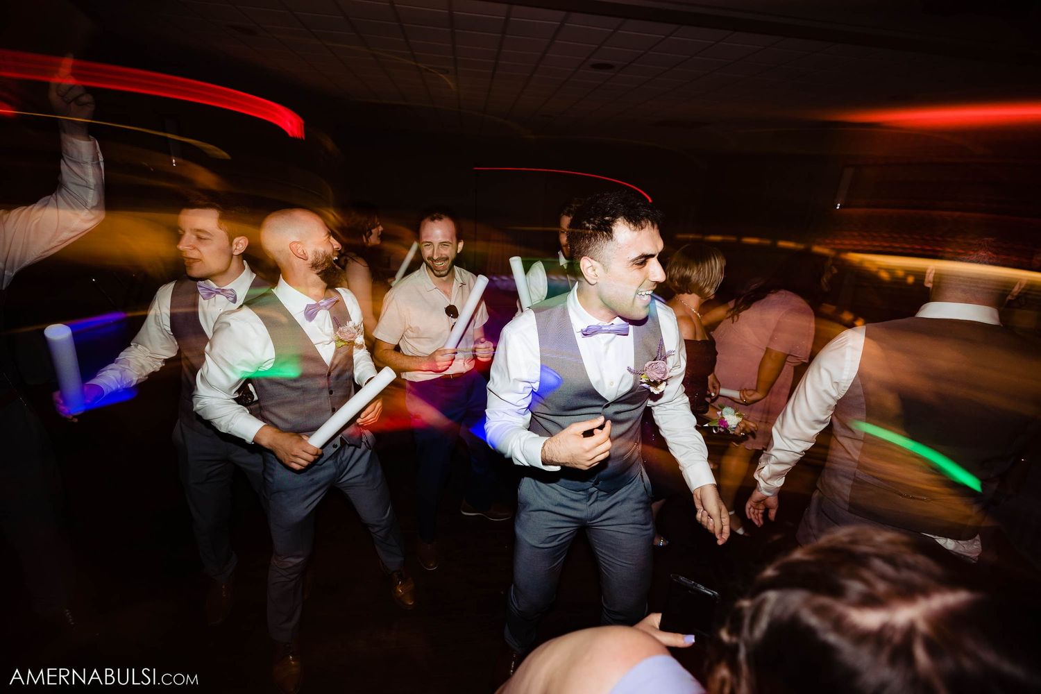 Wedding party dances with glow sticks at dimly lit indoor reception.