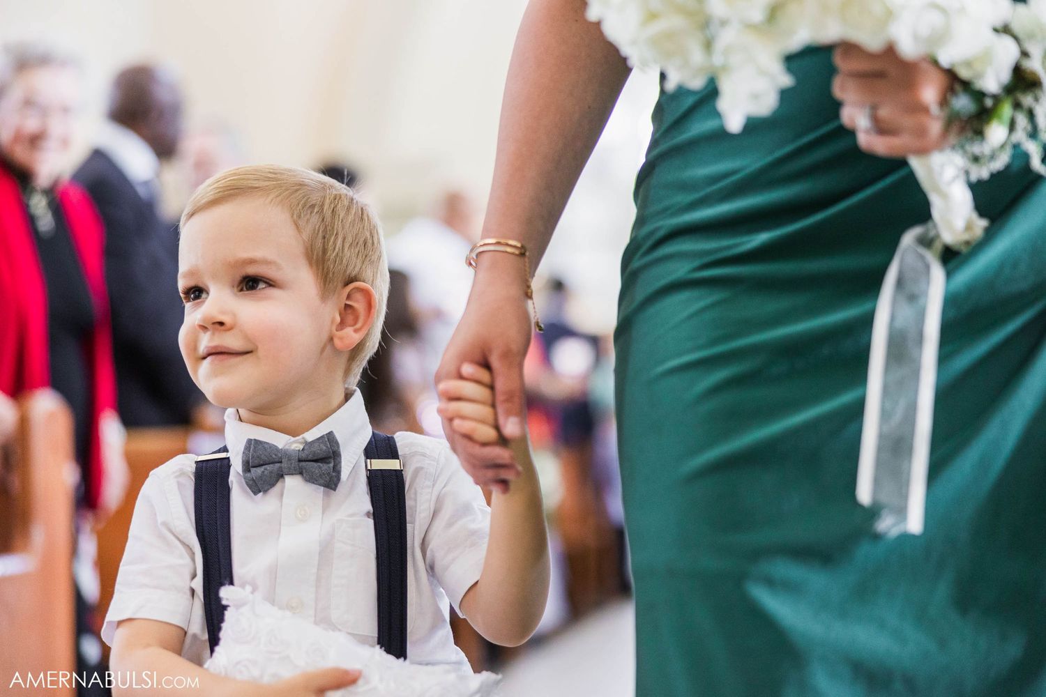 Young ring bearer in bow tie and suspenders stands next to emerald green bridesmaid dress.