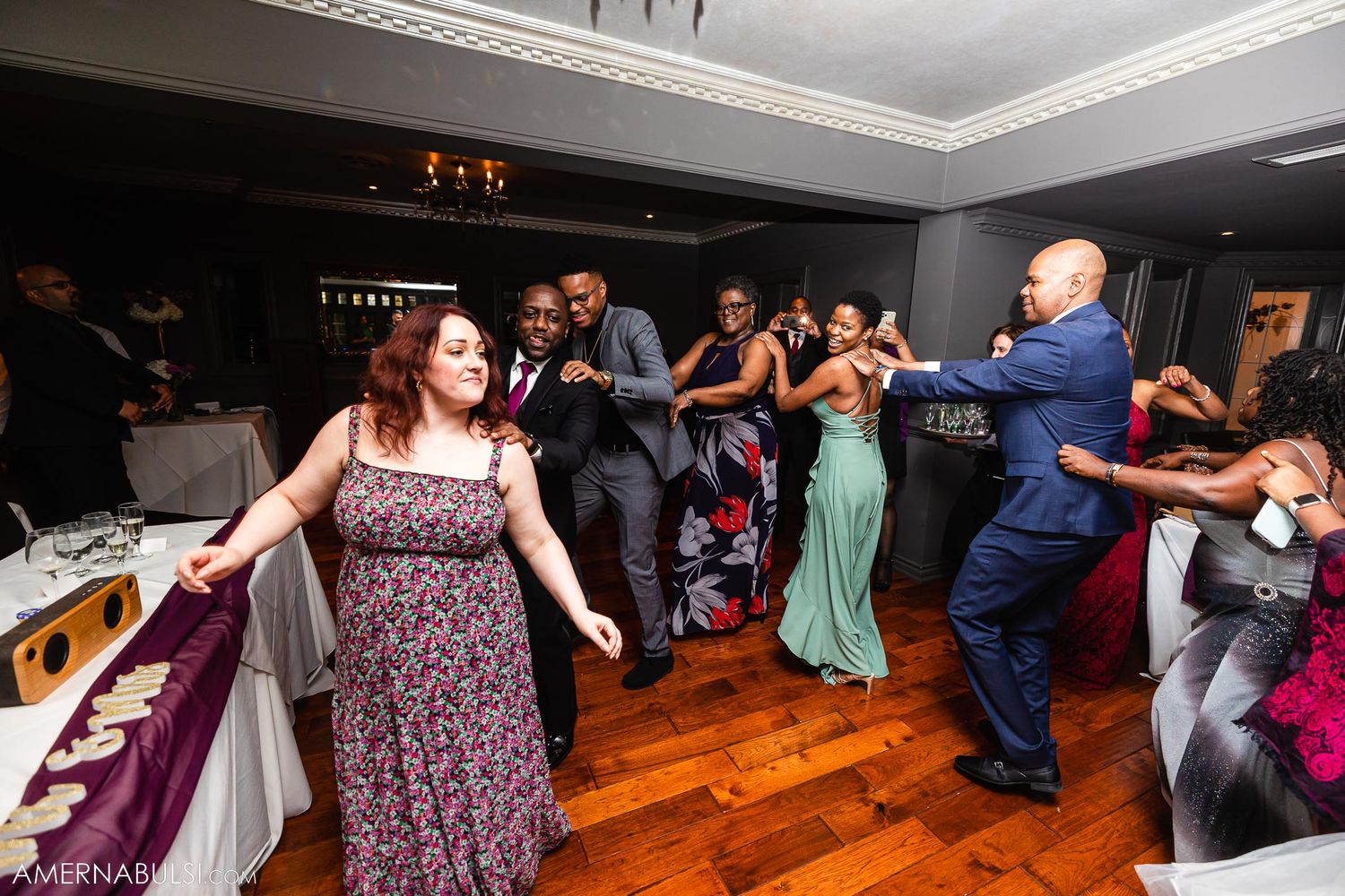 Wedding guests dancing in formation on wooden floor during reception celebration.