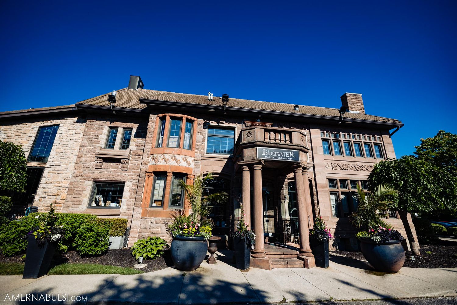 Stone mansion exterior with columns and large planters under blue sky.