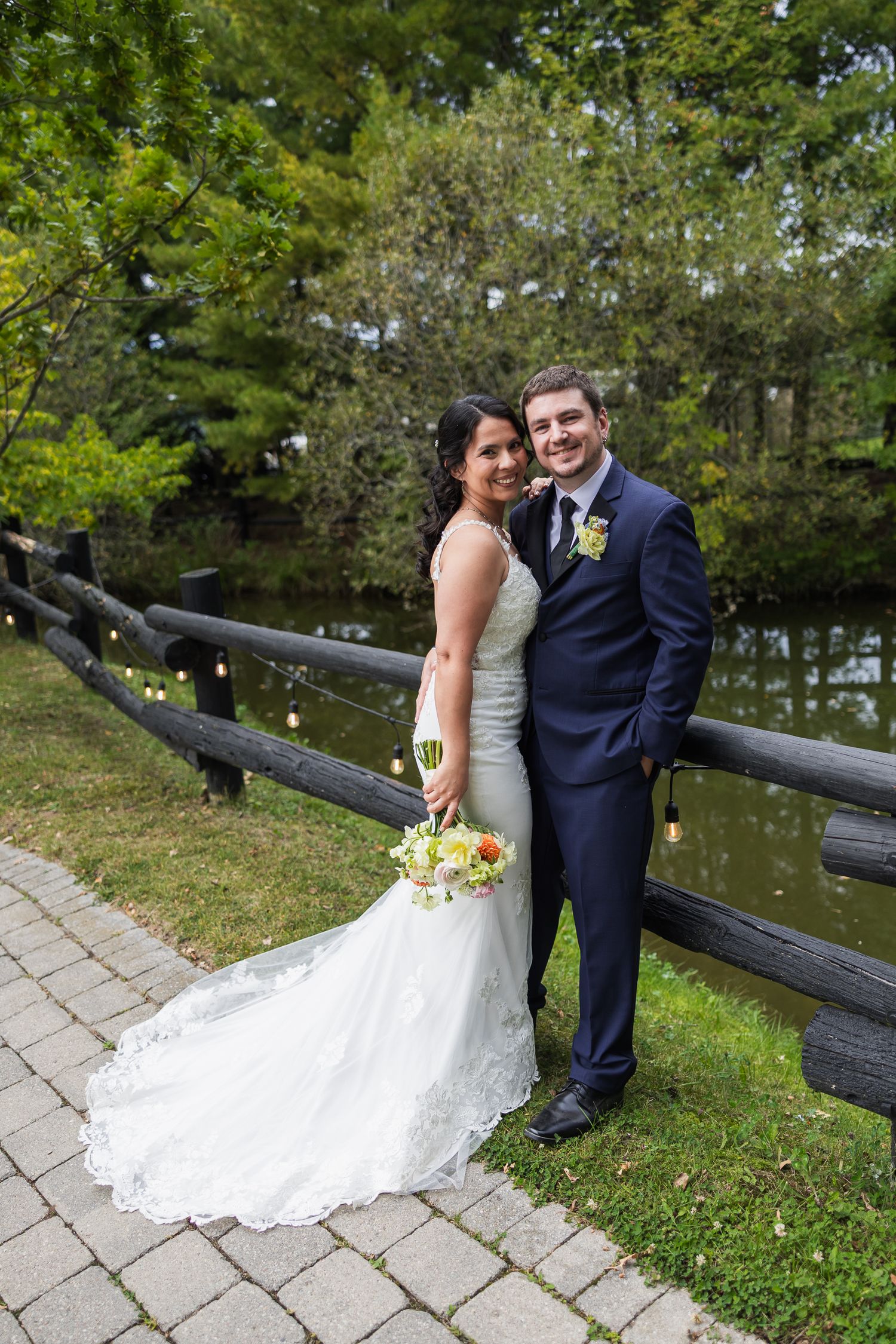 Bride and groom posing by a rustic wooden fence near a pond, surrounded by lush greenery on their wedding day.
