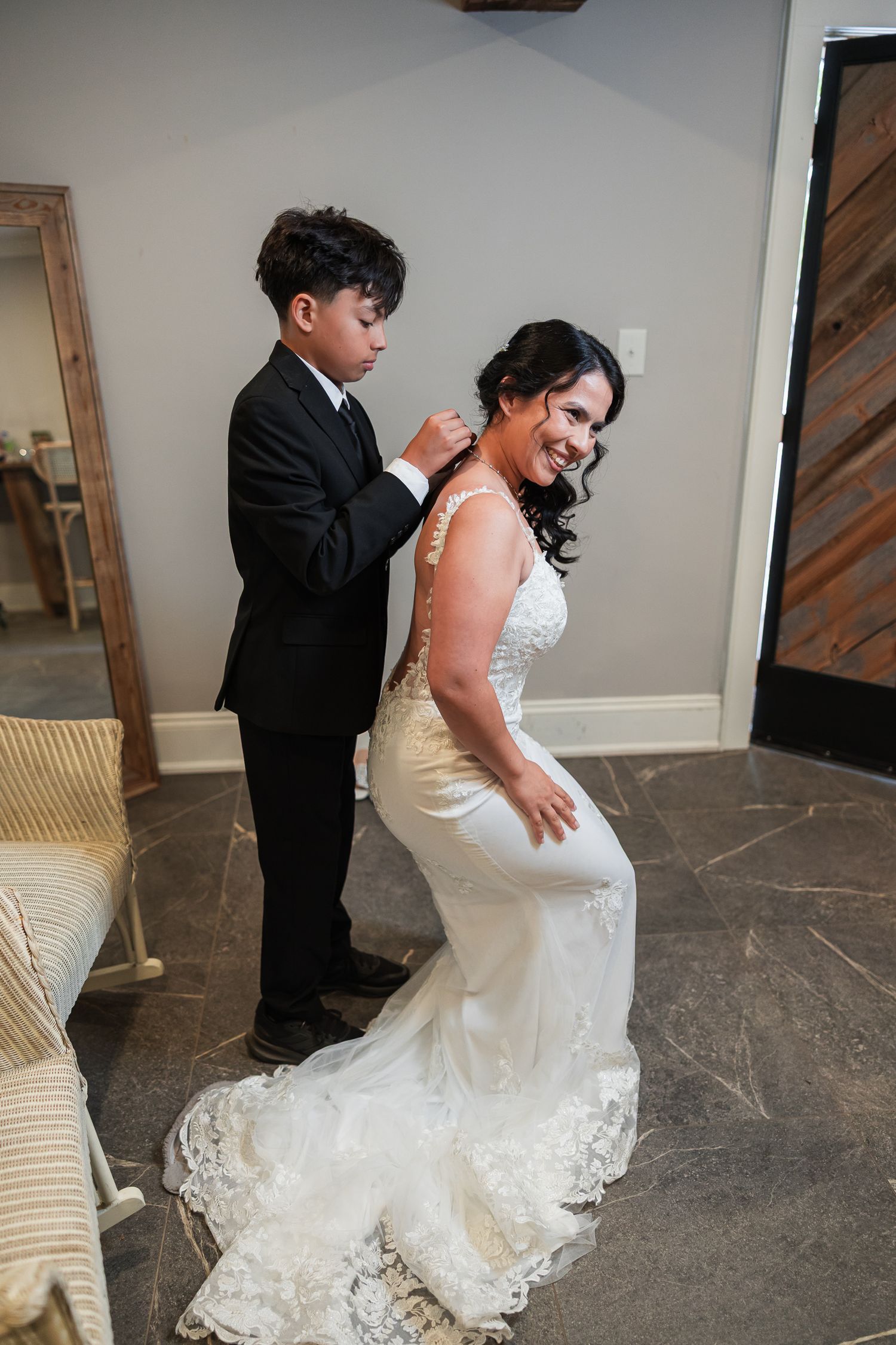 A smiling bride in a white lace gown gets her necklace fastened by a young boy in a black suit.