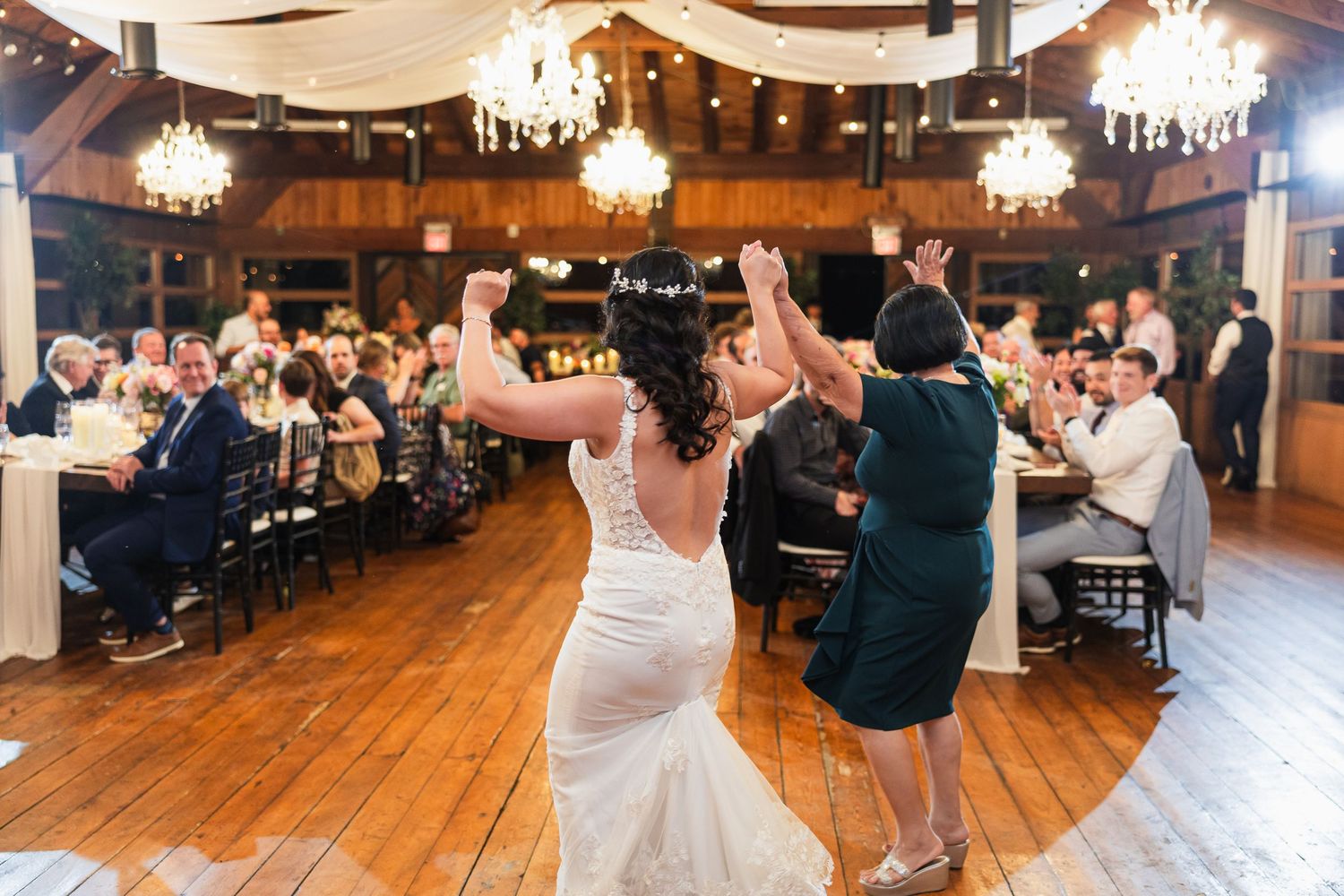 Bride and guest dancing at elegant rustic wedding reception with chandeliers and wood floors.