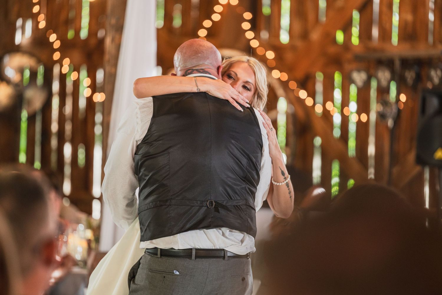 Bride and groom share an emotional first dance hug at rustic barn wedding reception with warm string lights.