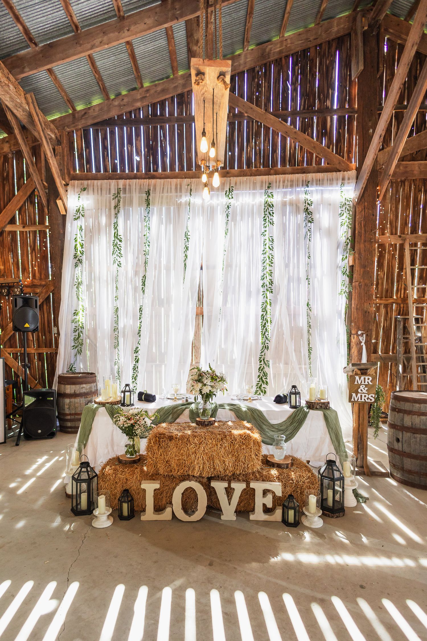 Rustic barn wedding sweetheart table decorated with hay bales, LOVE letters, lanterns, white drapes, and greenery.