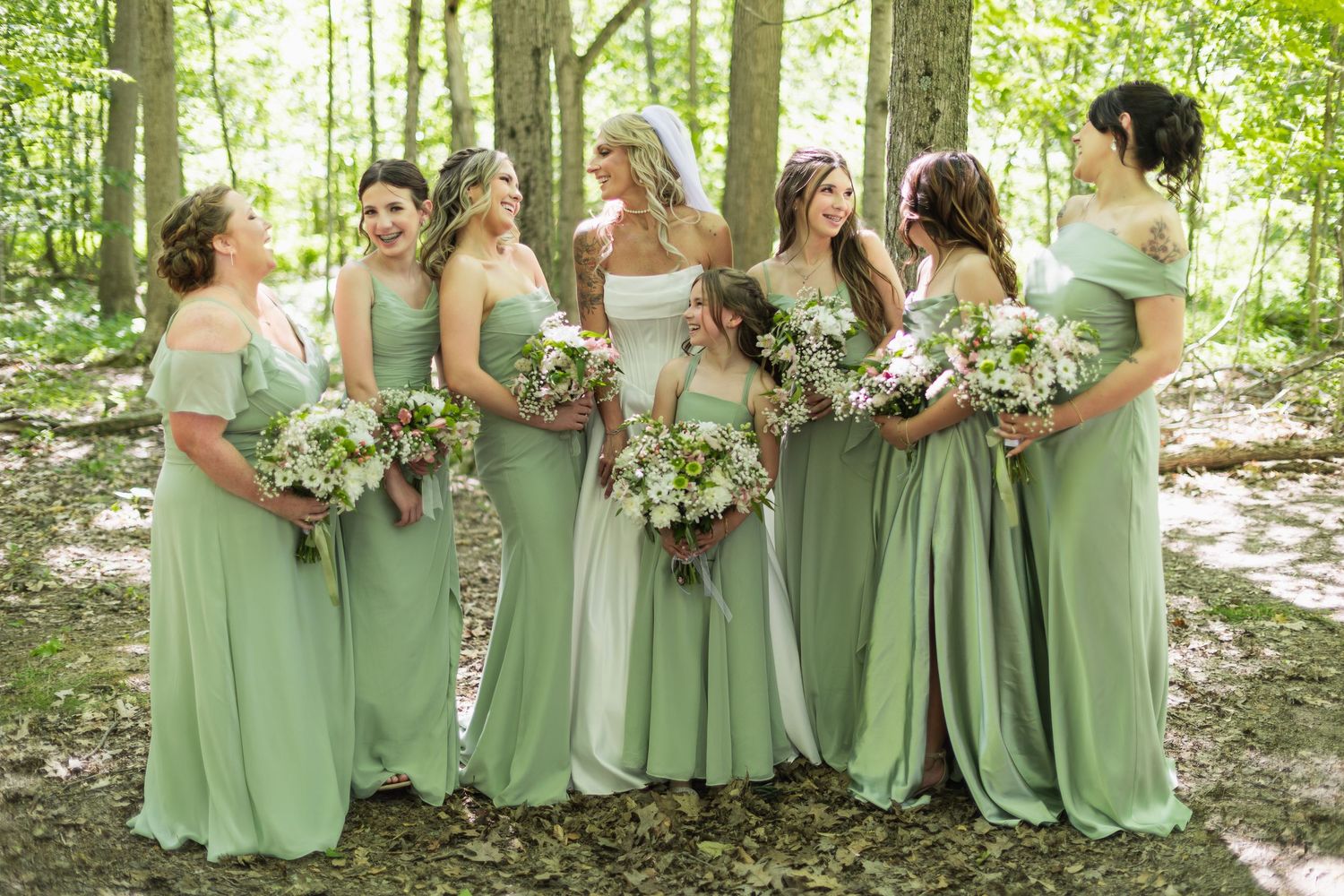 Bride and bridesmaids in sage green dresses holding white bouquets, smiling together in a wooded outdoor setting.