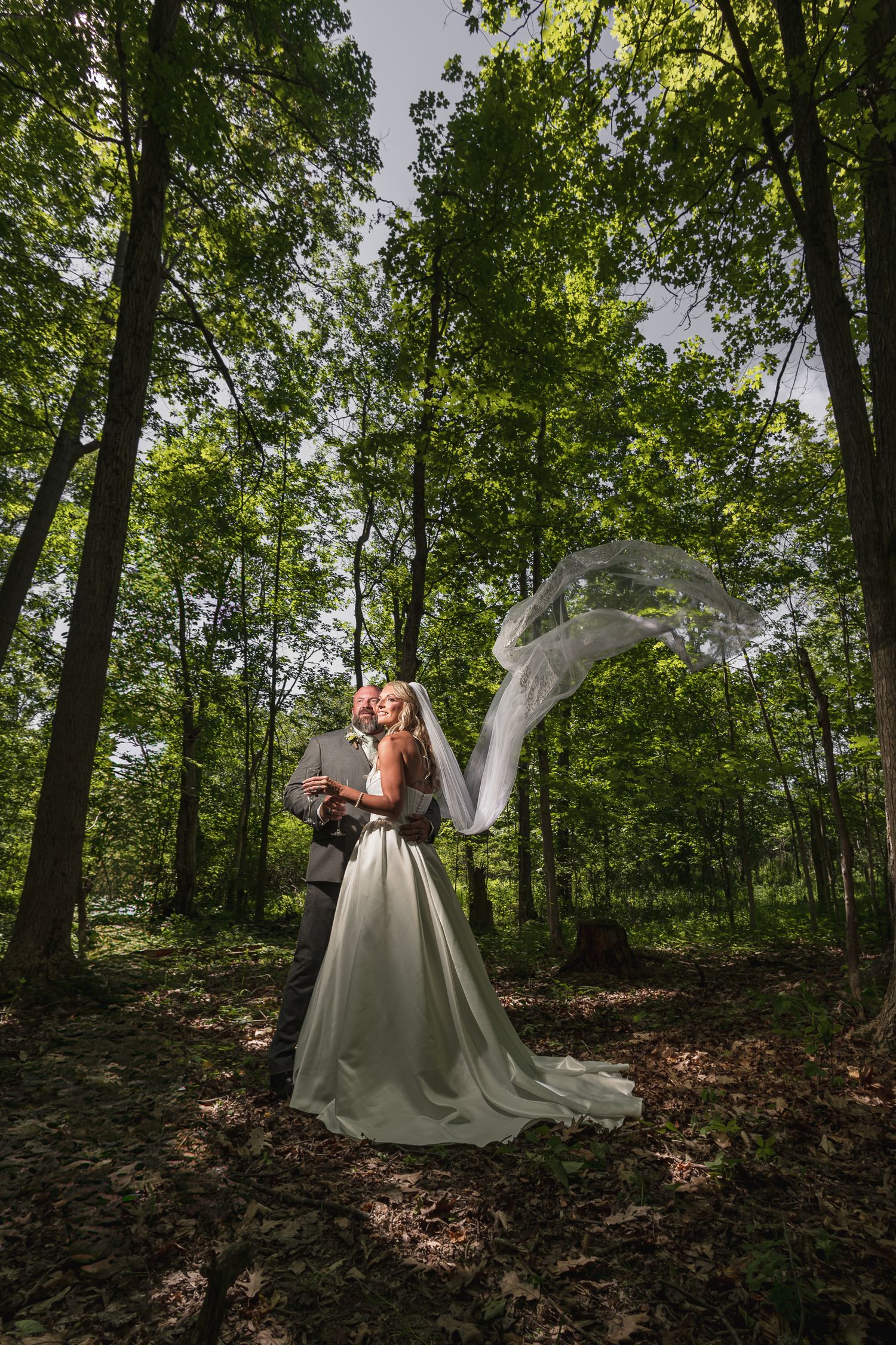 Bride and groom embrace in lush green forest with wedding veil flowing dramatically in the wind.