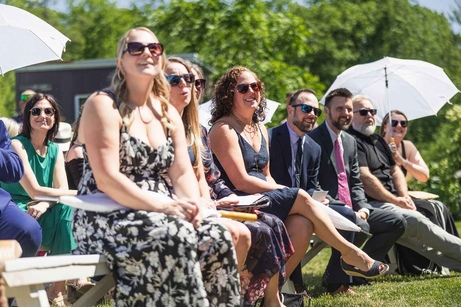 Smiling wedding guests seated outdoors in sunshine, some holding white parasols during ceremony.