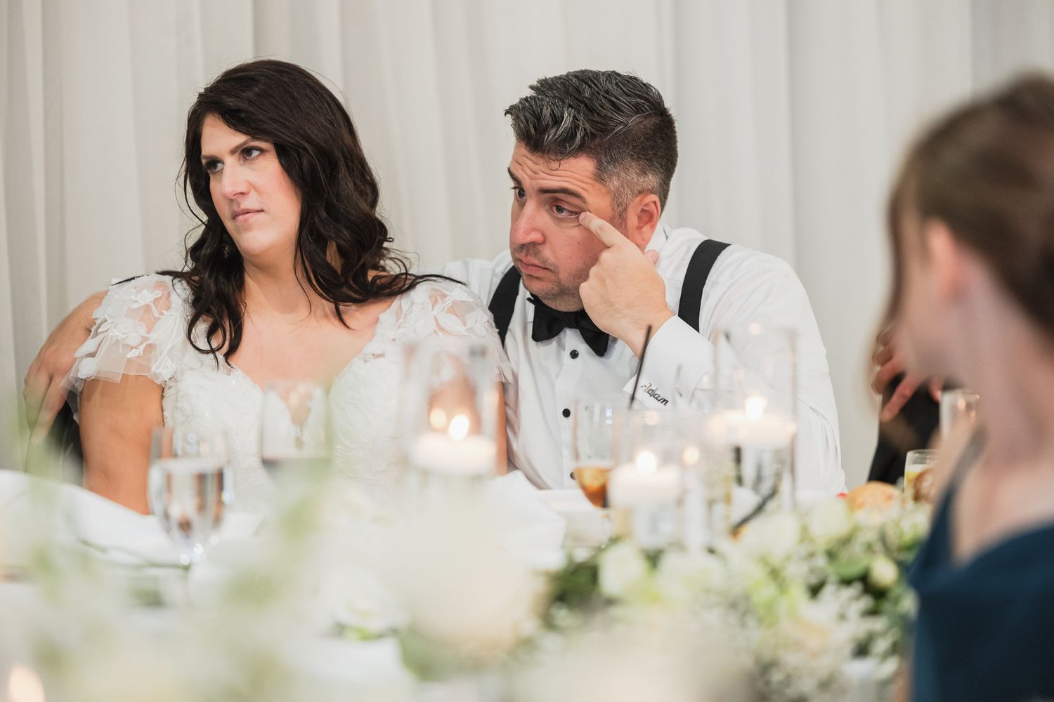 Bride and groom sitting at wedding reception head table listening to speeches with candles.