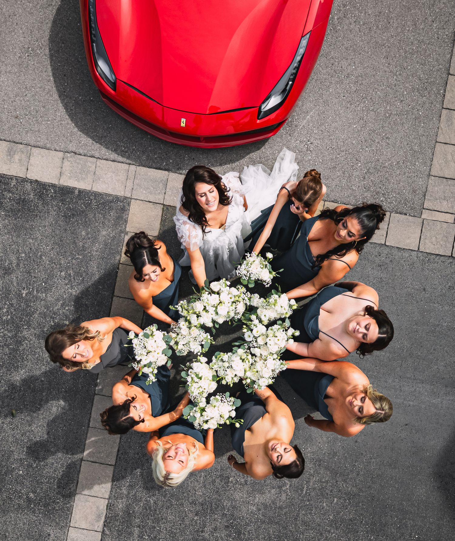 Aerial view of a bride and bridesmaids in navy dresses holding white bouquets, posing in front of a red Ferrari.