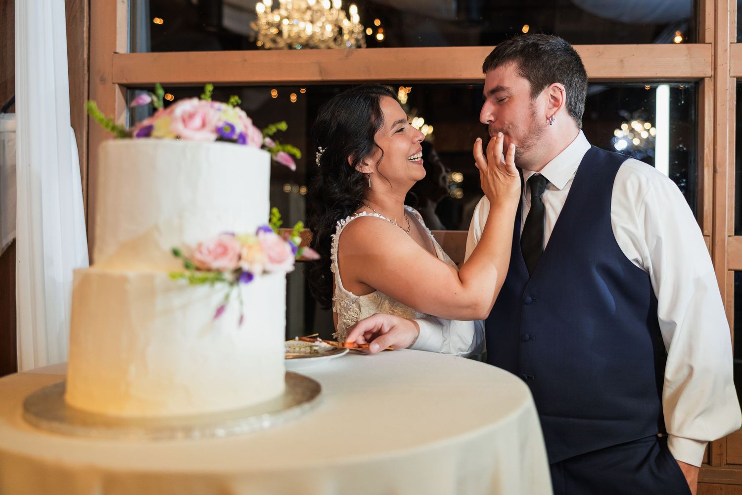 Bride feeds groom wedding cake at elegant reception with white floral cake on display.