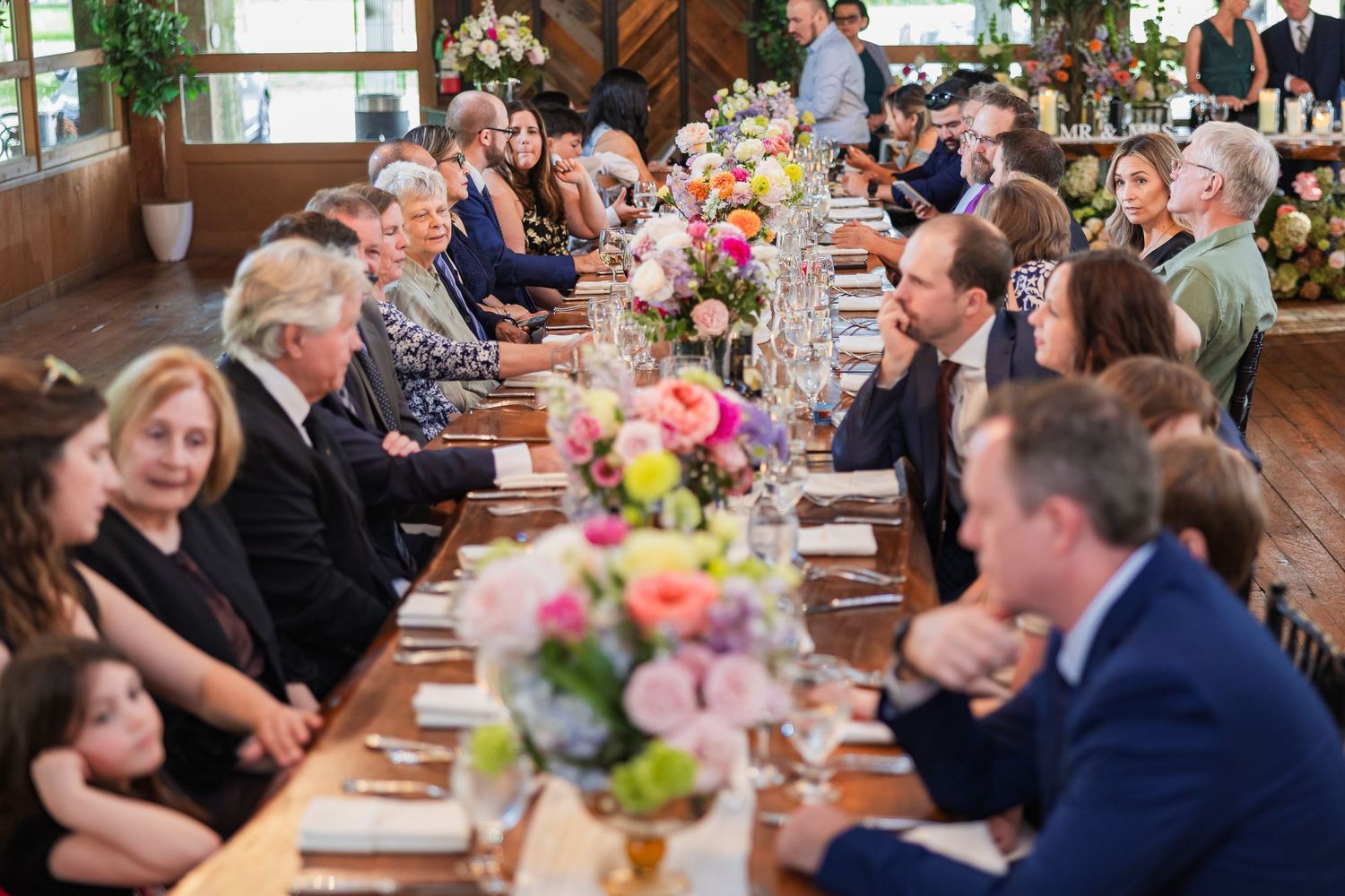 Wedding guests seated at long decorated reception tables with colorful floral centerpieces in a rustic barn venue.