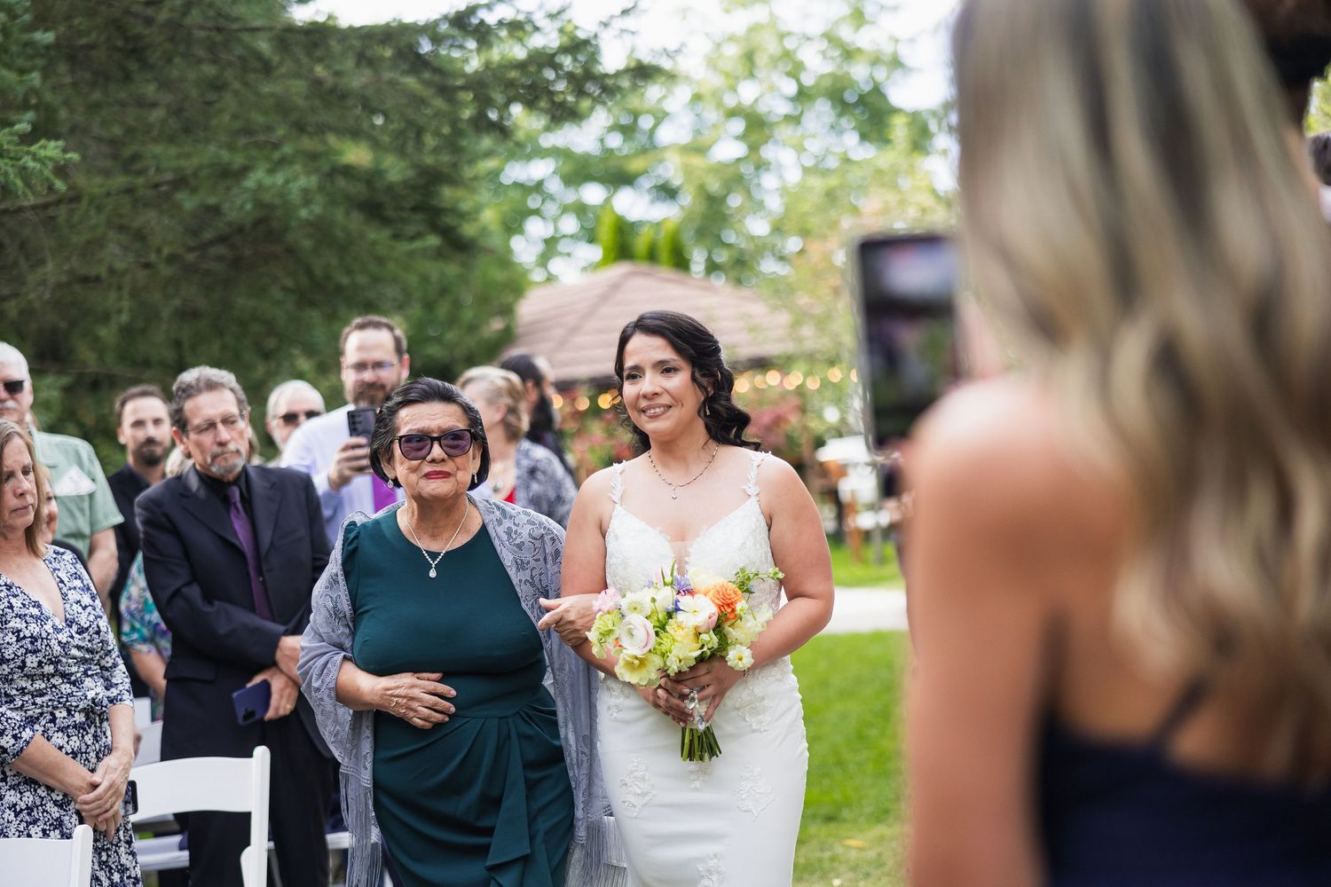Bride walks down the aisle with a woman in green, holding a colorful bouquet at outdoor wedding.