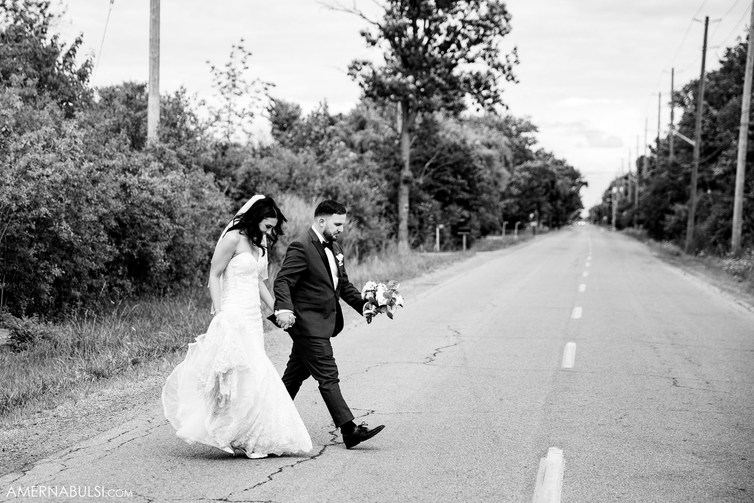 A black and white photo of newlyweds walking down a tree-lined road together.