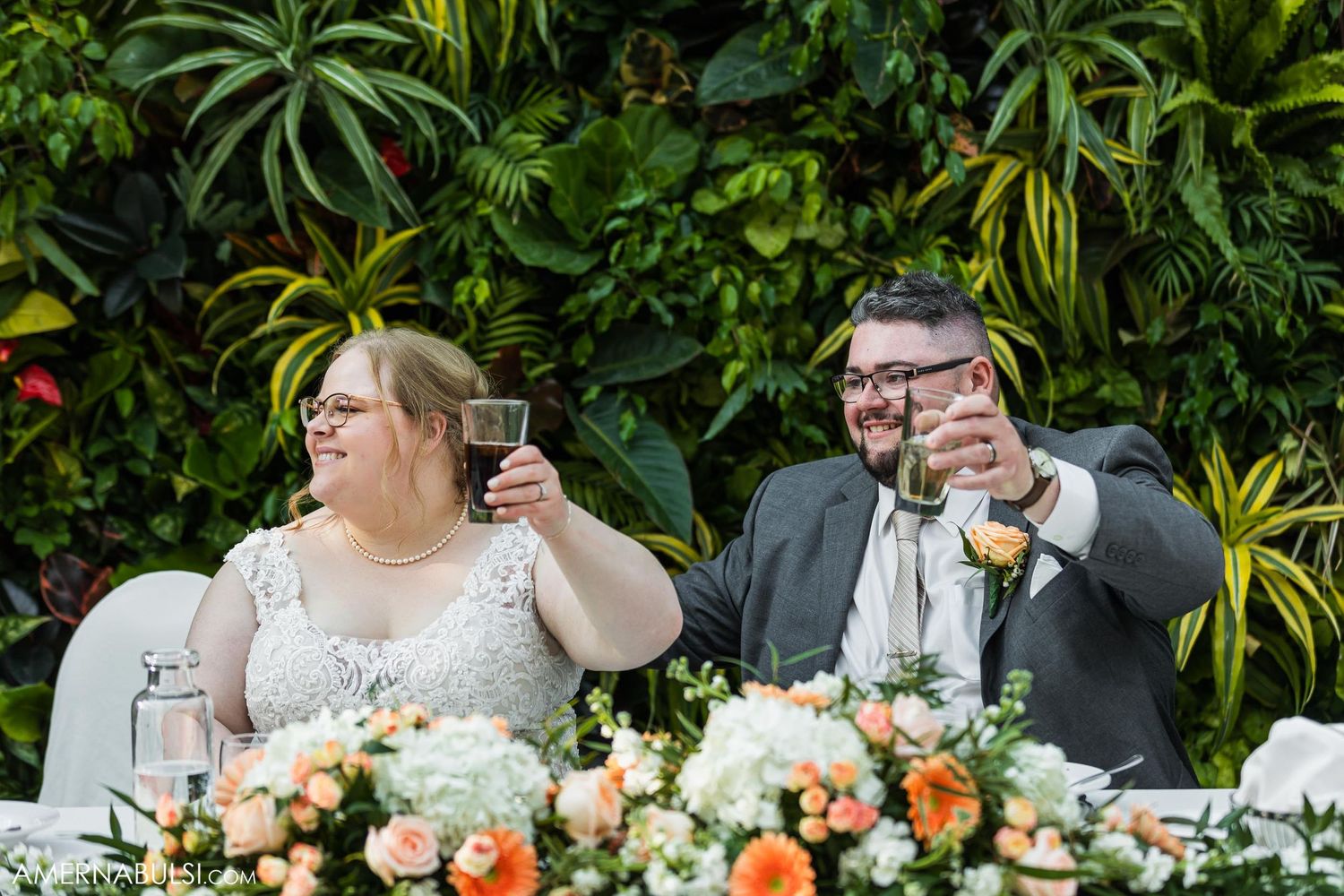 Newlyweds raise their glasses in a toast at their reception with tropical foliage backdrop.