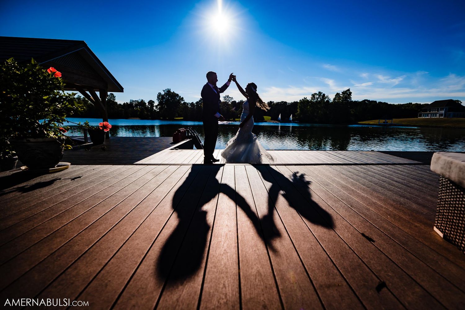 Dramatic silhouette of wedding couple dancing on dock against bright sunlit sky and lake.