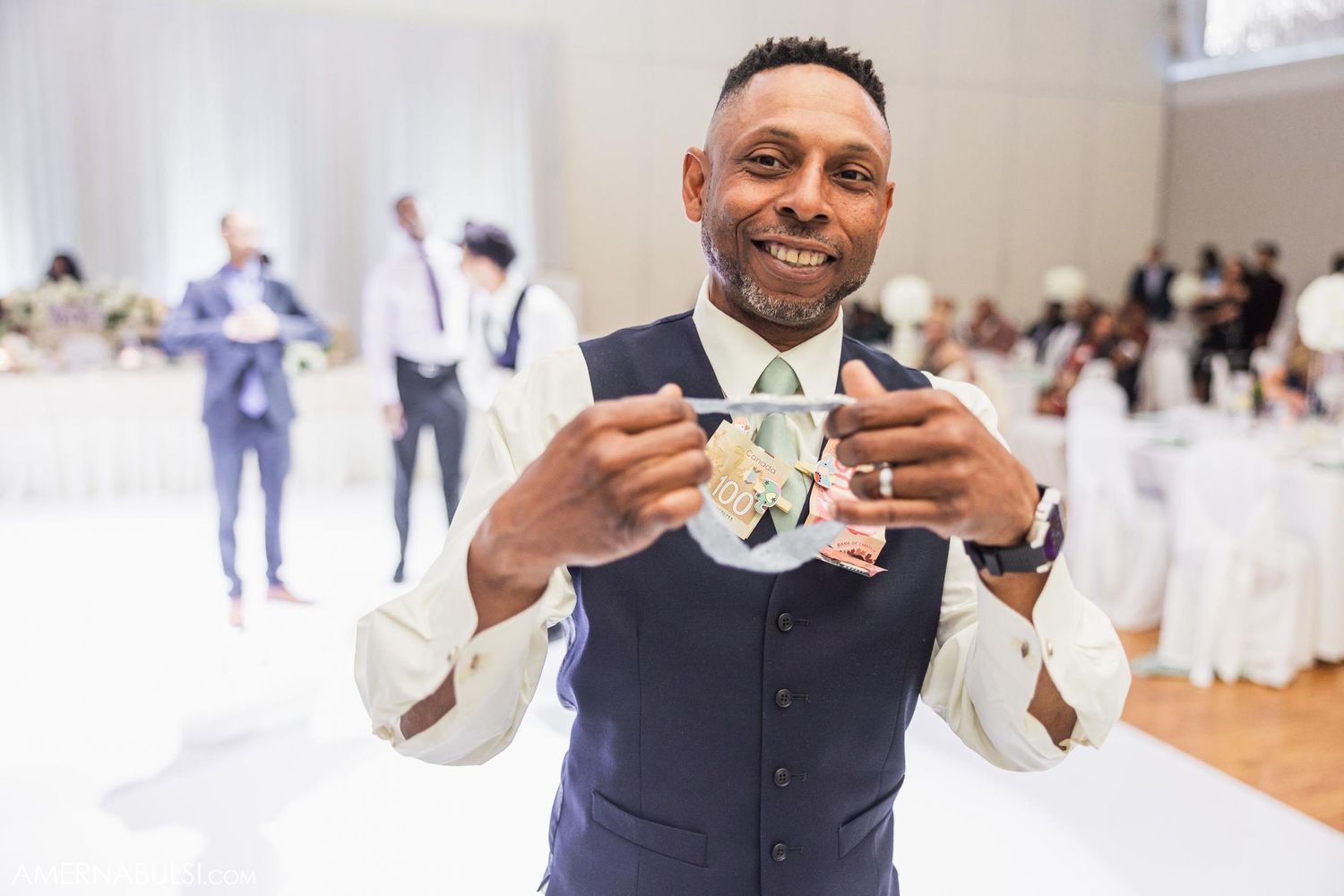 A groom in a navy vest poses with a playful smile while holding up his bowtie.