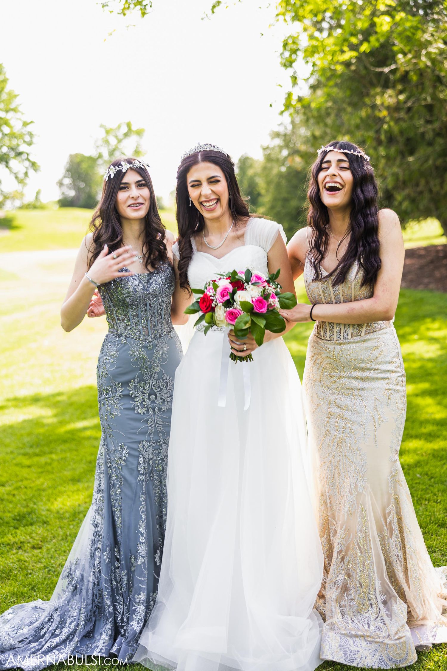 Three bridesmaids in elegant dresses laughing together outdoors while holding pink and white floral bouquet.
