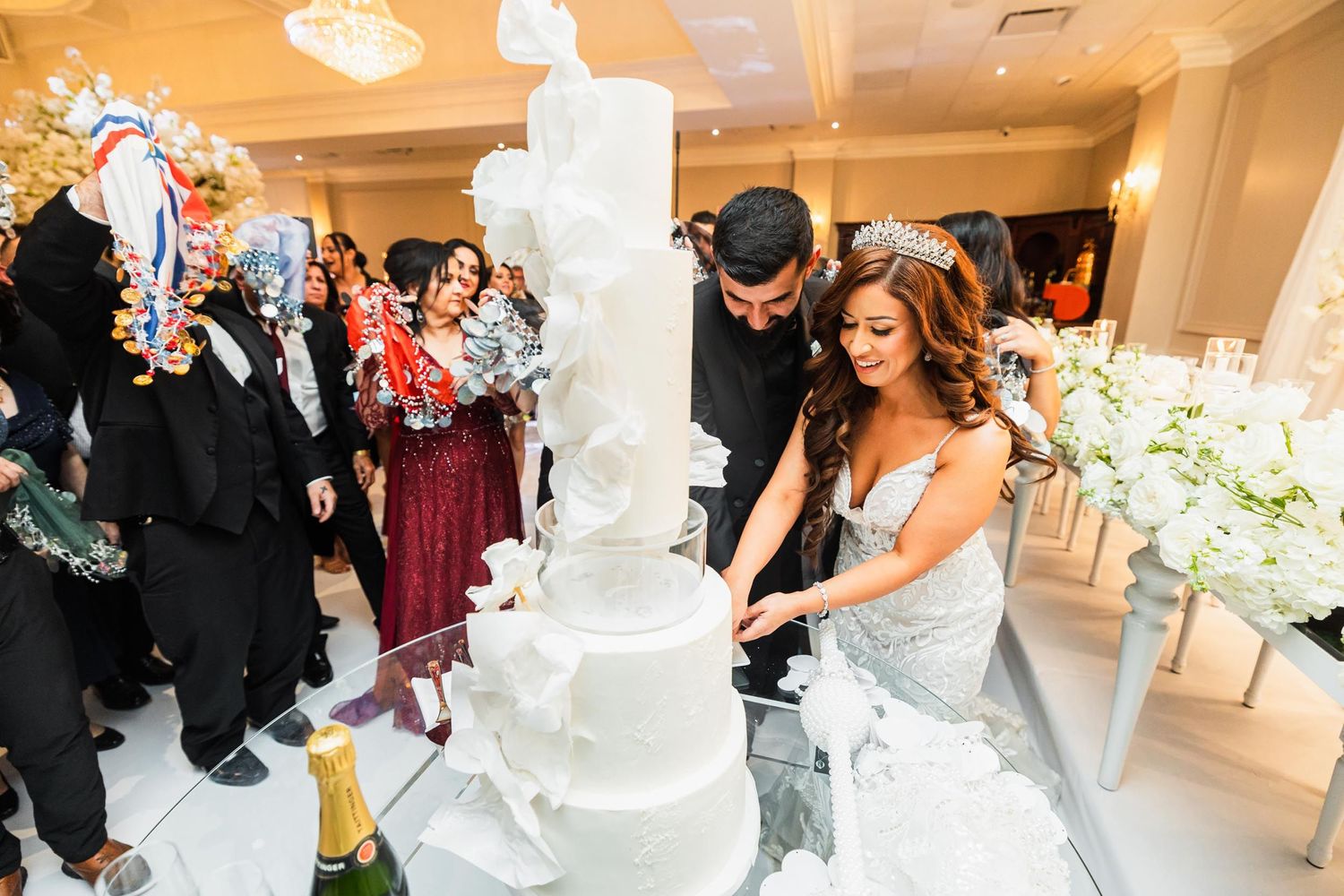 Couple cutting their elegant white tiered wedding cake surrounded by celebrating guests.